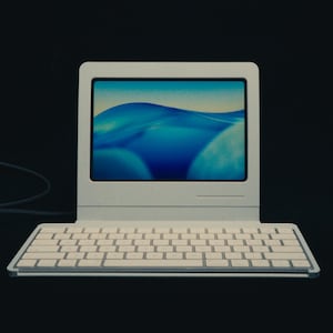 May include: A vintage-style white laptop with a keyboard and a screen displaying a blue and white abstract image. The laptop is set against a dark background, with a cable extending from the side.