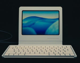 Peut inclure: Un ordinateur portable blanc de style rétro avec un clavier et un écran affichant une image abstraite bleue et blanche. L'ordinateur est posé sur un fond sombre, avec un câble qui s'étend sur le côté.