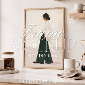 Op de afbeelding: Een ingelijste kunstdruk met een vrouw in een wit overhemd en een groene broek. De lijst is lichtbruin. De afbeelding maakt deel uit van een mockup met de tekst "Frame Mockup" en "ISO A DIN Ratio". Andere items zijn een koffiezetapparaat, mokken en een pot koffiebonen.