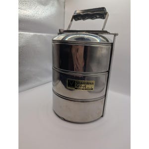 Könnte beinhalten: Ein Edelstahl-Tiffin-Träger mit drei gestapelten Fächern. Er hat einen Metallgriff mit schwarzem Griff. Der Träger hat ein Etikett mit der Aufschrift "Stainless Steel 18/8".