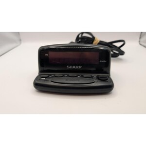 May include: A black Sharp digital alarm clock with a rectangular display screen, a prominent snooze button, and several smaller control buttons on the front panel. The device includes a black power cord extending from the back of the unit.