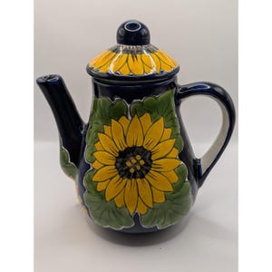 May include: A dark blue ceramic teapot featuring a hand-painted yellow sunflower design with green leaves. The teapot includes a matching lid with a yellow sunflower pattern and a dark blue knob. The spout and handle are dark blue.
