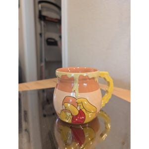 May include: A ceramic mug featuring Winnie the Pooh. The mug has a yellow handle and is decorated with a cartoon image of Winnie the Pooh in a red shirt. The mug has a honey pot design with dripping honey accents.