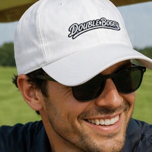 May include: White baseball cap with the text "DOUBLE BOGEY" in black script. The cap is worn by a person wearing sunglasses. The background is a green field.