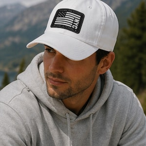 May include: A white baseball cap with a curved brim. The cap features a black rectangular patch with a stylized American flag and the word "NYC". The hat is made of a soft material and is suitable for outdoor activities.