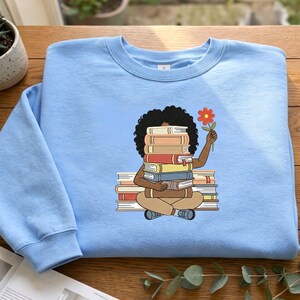 May include: A light blue crewneck sweatshirt featuring a graphic illustration of a person with dark curly hair sitting cross-legged while holding a stack of books in front of their face and a single orange flower in their raised hand.
