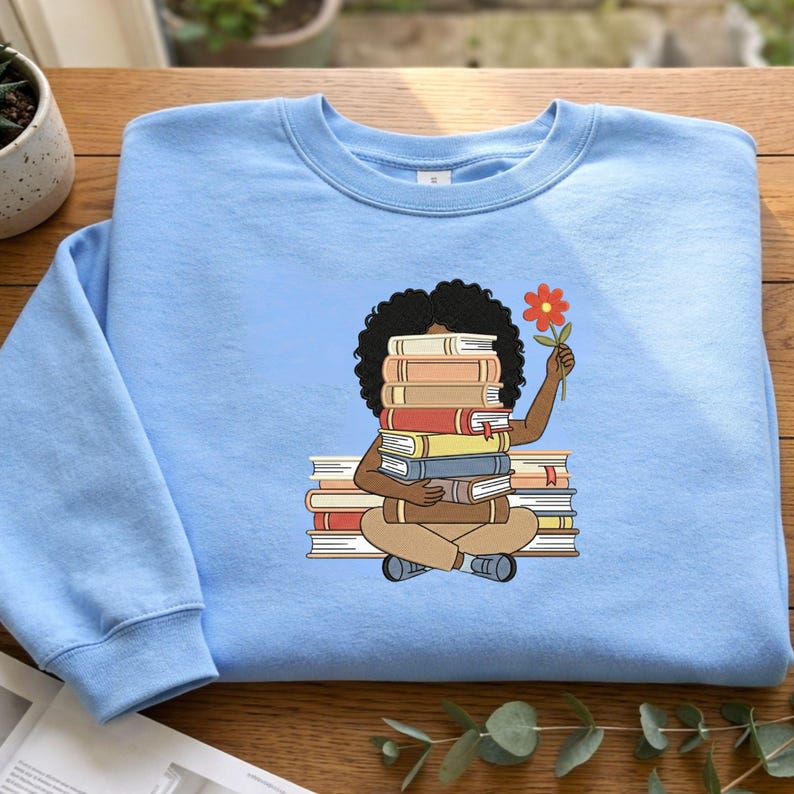 May include: A light blue crewneck sweatshirt featuring a graphic illustration of a person with dark curly hair sitting cross-legged while holding a stack of books in front of their face and a single orange flower in their raised hand.