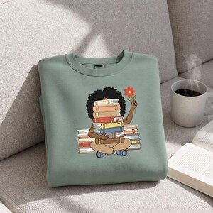May include: A sage green crewneck sweatshirt featuring a graphic illustration of a person with dark curly hair sitting cross-legged while holding a stack of books in front of their face and a single orange flower in their raised hand.