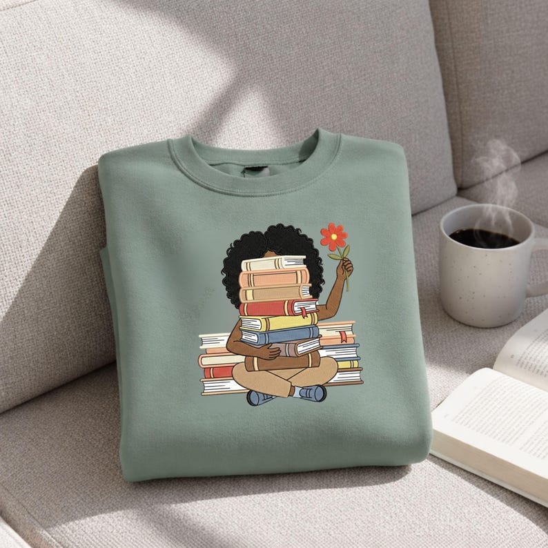 May include: A sage green crewneck sweatshirt featuring a graphic illustration of a person with dark curly hair sitting cross-legged while holding a stack of books in front of their face and a single orange flower in their raised hand.