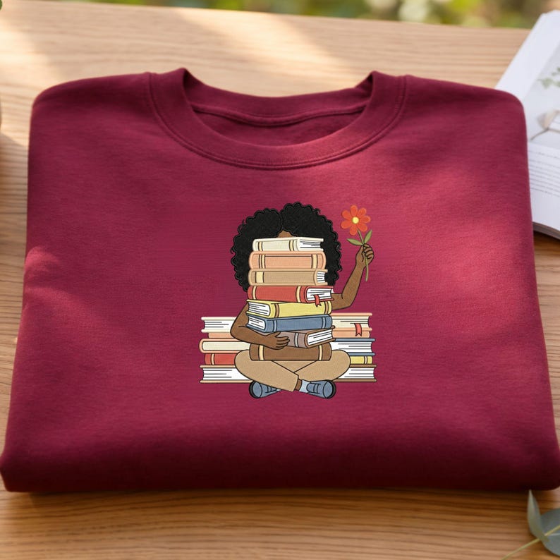 May include: A maroon crewneck sweatshirt featuring a graphic illustration of a person with dark curly hair sitting cross-legged, holding a stack of books that covers their face, and holding a single orange flower in their raised hand.