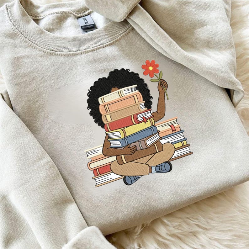 May include: A beige crewneck sweatshirt featuring a graphic print of a person with dark curly hair sitting cross-legged while holding a stack of books in front of their face and a single orange flower in their raised hand, wearing tan trousers.