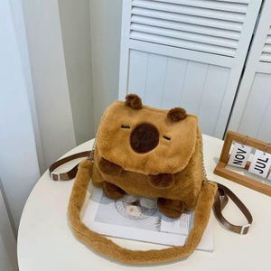 K&ouml;nnte beinhalten: Eine braune Pl&uuml;sch-Umh&auml;ngetasche im Capybara-Design mit weichem Kunstfell, kleinen abgerundeten Ohren, einem dunkelbraunen, kreisf&ouml;rmigen Nasenfleck und einem langen, verstellbaren Riemen mit metallischen Kettenakzenten an den Seiten.