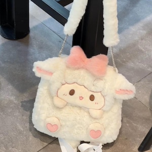 K&ouml;nnte beinhalten: Eine wei&szlig;e Pl&uuml;sch-Umh&auml;ngetasche in Schaf-Form mit weicher Textur. Die Tasche hat eine rosa Schleife oben, zwei rosa gef&uuml;tterte Ohren und ein Cartoon-Gesicht mit braunen Augen. Sie verf&uuml;gt &uuml;ber einen langen wei&szlig;en Kunstfellriemen und zwei kleine rosa Herzf&uuml;&szlig;e.