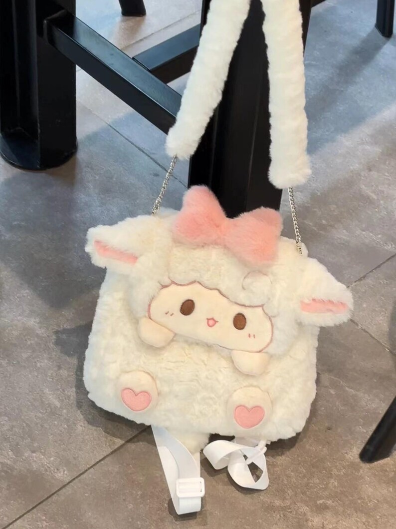 K&ouml;nnte beinhalten: Eine wei&szlig;e Pl&uuml;sch-Umh&auml;ngetasche in Schaf-Form mit weicher Textur. Die Tasche hat eine rosa Schleife oben, zwei rosa gef&uuml;tterte Ohren und ein Cartoon-Gesicht mit braunen Augen. Sie verf&uuml;gt &uuml;ber einen langen wei&szlig;en Kunstfellriemen und zwei kleine rosa Herzf&uuml;&szlig;e.