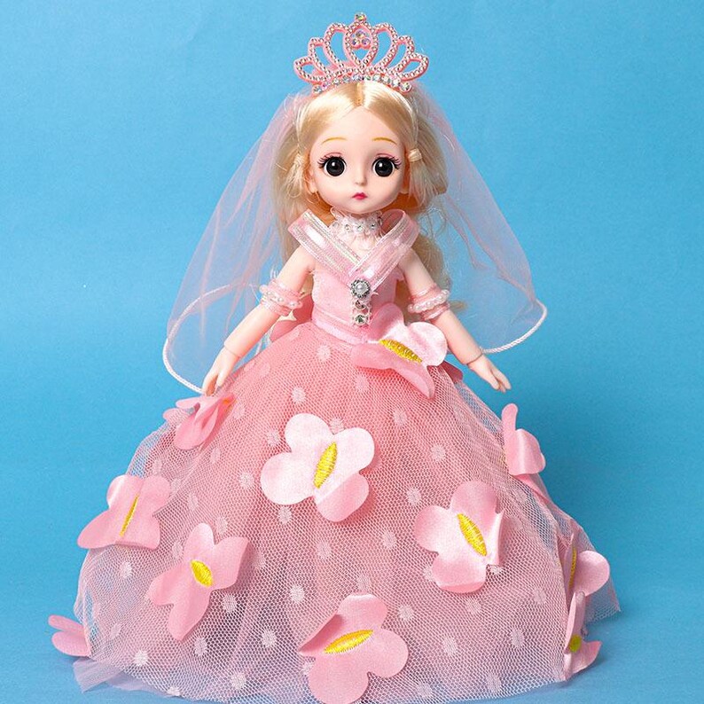Puede incluir: Una mu&ntilde;eca de cabello rubio viste un vestido de tul rosa adornado con flores de tela rosa con centros amarillos. La mu&ntilde;eca lleva una tiara rosa con cristales transparentes y un velo a juego. El corpi&ntilde;o incluye un adorno de tono plateado.