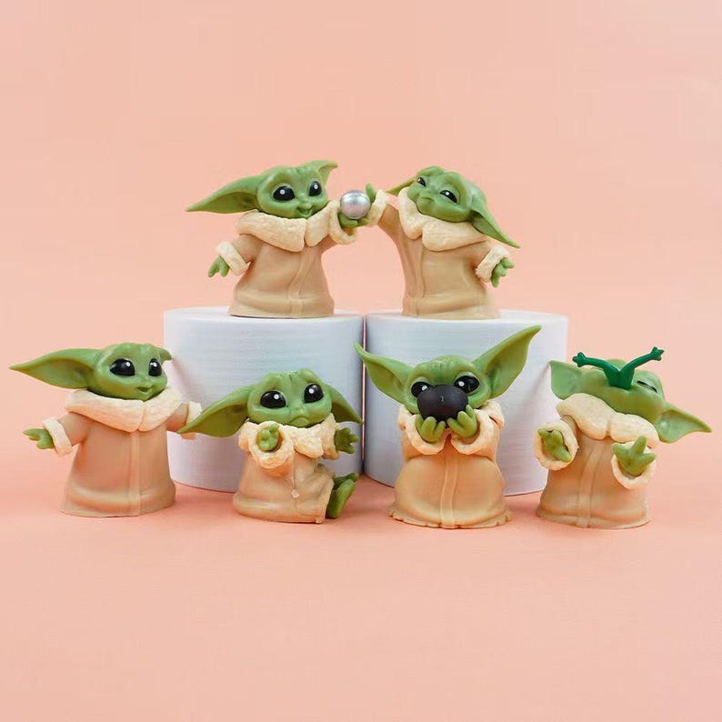 May include: A set of six small green alien figurines with large black eyes and pointed ears, each wearing a tan robe with a cream-colored collar. The figures are posed in various positions, with some holding small round objects or interacting with each other.