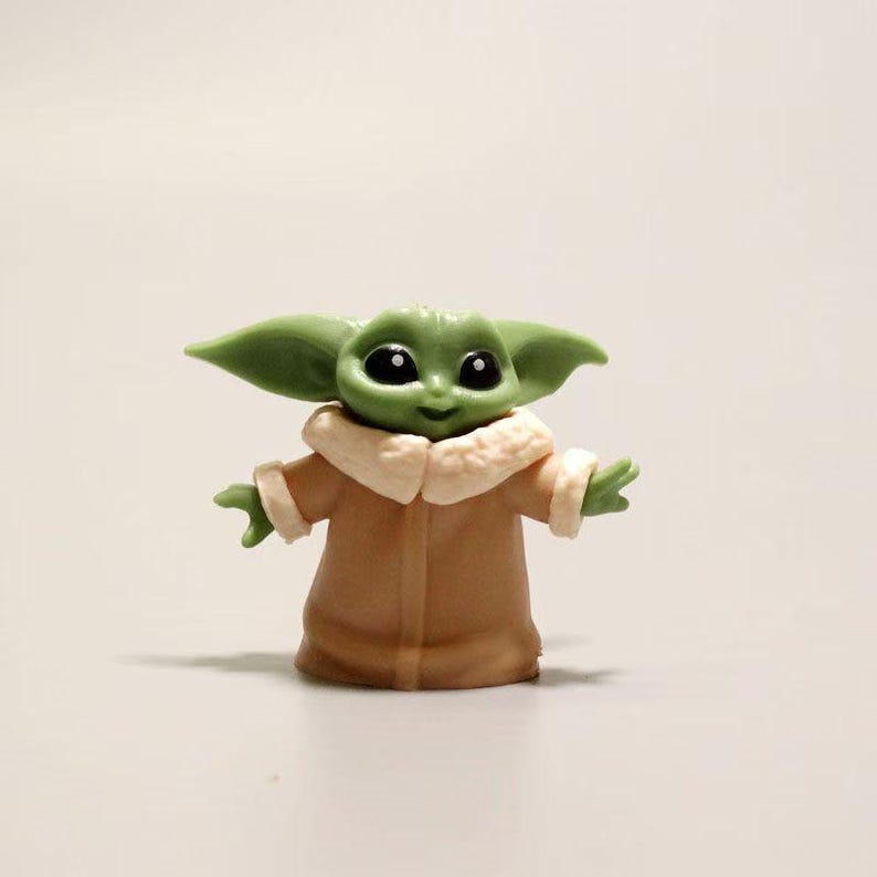 May include: A small green figurine with large pointed ears and wide black eyes. The character wears a tan robe featuring a thick, textured cream-colored collar and matching cuffs. The figure stands in a neutral pose with arms slightly extended.
