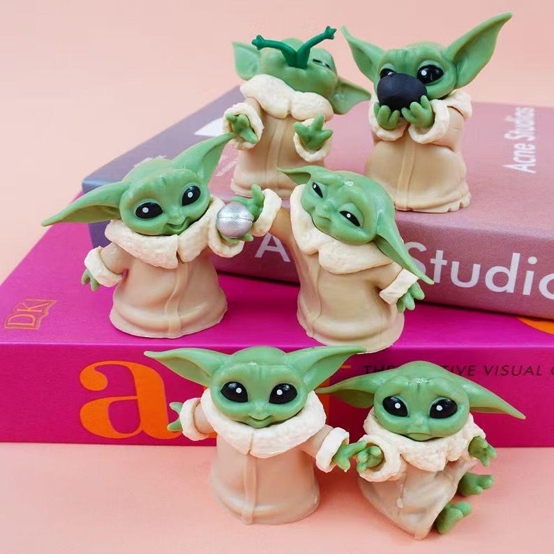May include: Six small green figurines with large ears and black eyes, each wearing a tan robe with a white collar. One figurine holds a silver sphere, another holds a black sphere, and one has green antennae. They are arranged on two stacked books.