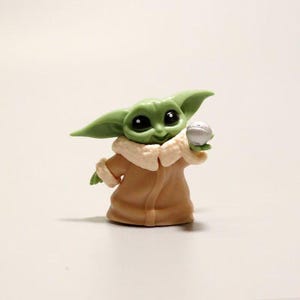 May include: A small green figurine of a creature with large dark eyes and pointed ears, wearing a tan robe with a cream-colored collar. The figure is holding a small, round, silver-colored ball in one hand, featuring a smooth, matte finish.