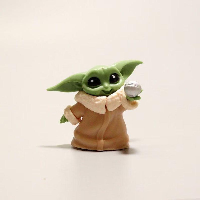 May include: A small green figurine of a creature with large dark eyes and pointed ears, wearing a tan robe with a cream-colored collar. The figure is holding a small, round, silver-colored ball in one hand, featuring a smooth, matte finish.