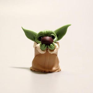 May include: A small green creature figurine with large pointed ears and wide dark eyes, wearing a tan robe with a cream-colored collar. The figure is holding a small dark brown spherical object with both hands near its face.