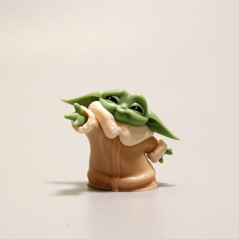 May include: A small green creature figurine with large pointed ears and dark eyes, wearing a tan robe with a wide, light-colored collar. The figure is posed with one arm extended forward, featuring three-fingered hands and a smooth, matte finish.