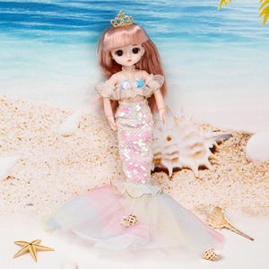 Puede incluir: Una mu&ntilde;eca con cabello largo rosa, una corona dorada, un top con volantes y hombros descubiertos con un detalle de concha azul, y una falda de sirena con lentejuelas que termina en una cola de tul pastel multicolor con una peque&ntilde;a concha.