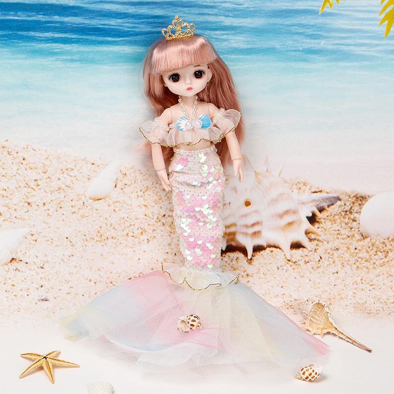 Puede incluir: Una mu&ntilde;eca con cabello largo rosa, una corona dorada, un top con volantes y hombros descubiertos con un detalle de concha azul, y una falda de sirena con lentejuelas que termina en una cola de tul pastel multicolor con una peque&ntilde;a concha.