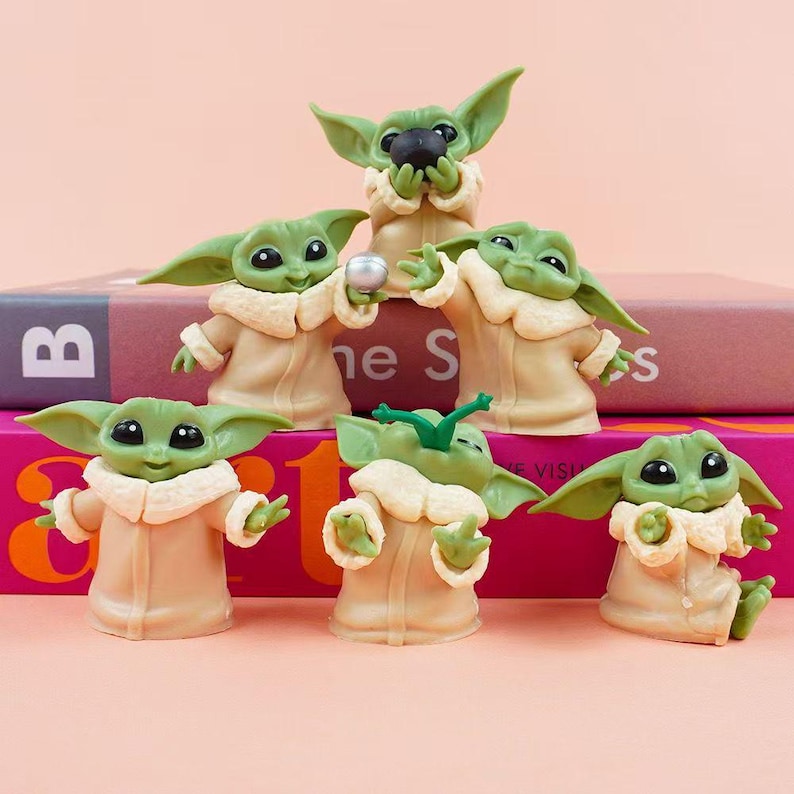 May include: A collection of six small green alien figurines with large black eyes and pointed ears, each wearing a tan robe with a cream-colored collar. The figures are posed in various actions, including holding a small black sphere or a silver ball.
