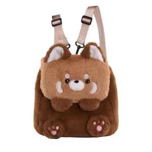 K&ouml;nnte beinhalten: Ein Pl&uuml;schrucksack in Form eines Roten Pandas mit weichem braunem Fell, wei&szlig;en Ohr-Akzenten und einem freundlichen Gesicht. Der Rucksack verf&uuml;gt &uuml;ber verstellbare beige Tr&auml;ger, eine kleine gestickte Nase und zwei Pfoten mit rosa Ballen.