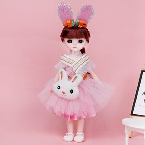 Puede incluir: Una mu&ntilde;eca de pelo casta&ntilde;o con un vestido de tul rosa, un chal blanco con perlas y un bolso de peluche blanco con forma de conejo. La mu&ntilde;eca lleva una diadema con orejas de conejo rosas brillantes y peque&ntilde;os detalles de zanahorias.