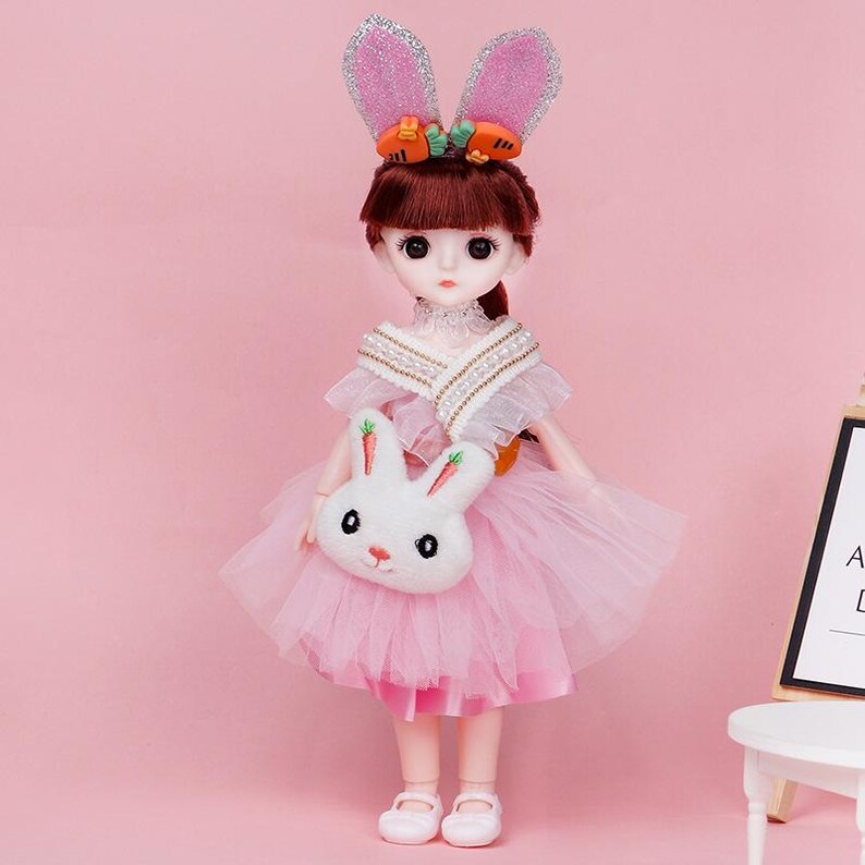 Puede incluir: Una mu&ntilde;eca de pelo casta&ntilde;o con un vestido de tul rosa, un chal blanco con perlas y un bolso de peluche blanco con forma de conejo. La mu&ntilde;eca lleva una diadema con orejas de conejo rosas brillantes y peque&ntilde;os detalles de zanahorias.