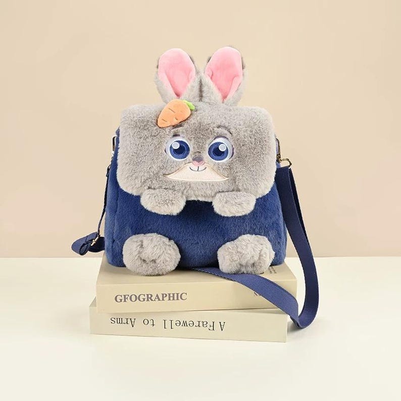 K&ouml;nnte beinhalten: Ein flauschiger Pl&uuml;schrucksack in Dunkelblau und Grau, geformt wie ein Hase mit gro&szlig;en blauen Augen, rosa gef&uuml;tterten Ohren und einer kleinen orangefarbenen Karotte. Die Tasche hat einen Klappverschluss, zwei graue Pfoten vorne und einen Schulterriemen.