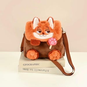 K&ouml;nnte beinhalten: Eine Pl&uuml;sch-Umh&auml;ngetasche in Fuchsform mit orangefarbenem und braunem Fell. Die Tasche zeigt ein Fuchskopf-Motiv auf der Klappe, zwei orangefarbene Pfoten am Boden und ein rosafarbenes Pfoten-Lutscher-Accessoire. Sie hat einen braunen Schulterriemen.