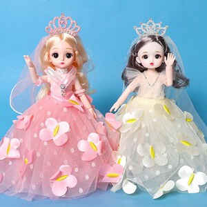 Puede incluir: Dos mu&ntilde;ecas con vestidos de gala y tiaras. La mu&ntilde;eca de la izquierda tiene cabello rubio y un vestido rosa con aplicaciones de mariposas rosas. La mu&ntilde;eca de la derecha tiene cabello oscuro y un vestido crema con aplicaciones de mariposas blancas.