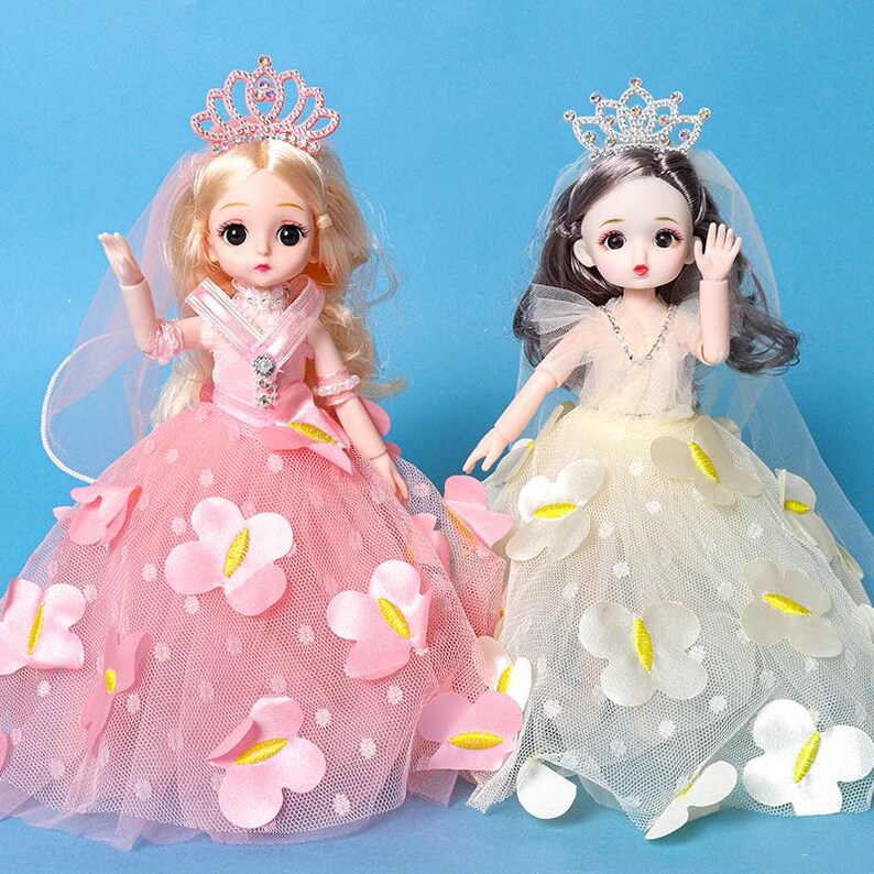 Puede incluir: Dos mu&ntilde;ecas con vestidos de gala y tiaras. La mu&ntilde;eca de la izquierda tiene cabello rubio y un vestido rosa con aplicaciones de mariposas rosas. La mu&ntilde;eca de la derecha tiene cabello oscuro y un vestido crema con aplicaciones de mariposas blancas.