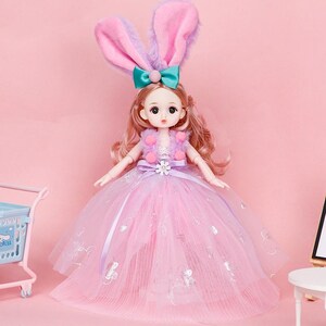 Puede incluir: Una mu&ntilde;eca con cabello largo casta&ntilde;o claro que viste un vestido de tul morado y rosa con una flor blanca en la cintura. La mu&ntilde;eca lleva un chaleco de peluche morado con pompones rosas y una diadema con orejas de conejo rosas y un lazo.
