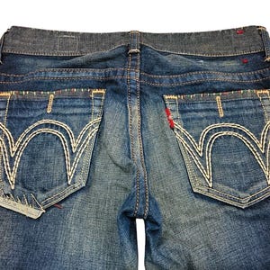 May include: Close-up of blue denim jeans. The jeans feature large back pockets with white decorative stitching. The waistband is a darker shade of blue. Red stitching accents the pockets and seams. The jeans are slightly faded.