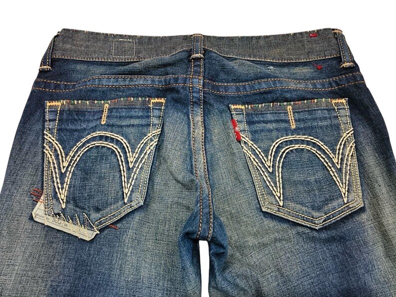 May include: Close-up of blue denim jeans. The jeans feature large back pockets with white decorative stitching. The waistband is a darker shade of blue. Red stitching accents the pockets and seams. The jeans are slightly faded.