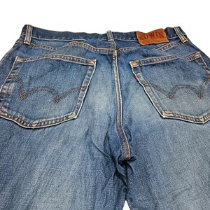 Puede incluir: Primer plano de unos vaqueros de mezclilla azules con dos bolsillos traseros con un dise&ntilde;o de costura distintivo. Los vaqueros tienen un parche de cuero marr&oacute;n con la marca "EDWIN" y detalles de costura rojos. Los vaqueros son de estilo cl&aacute;sico.
