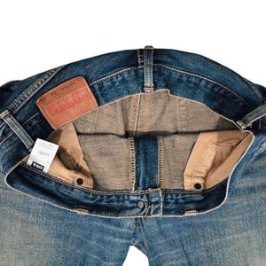 May include: Close-up of blue denim jeans, showing the inside details. The jeans have a faded wash, a brown leather patch, and a white tag. The jeans are unbuttoned, revealing the interior construction and pockets.