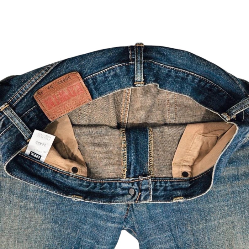 May include: Close-up of blue denim jeans, showing the inside details. The jeans have a faded wash, a brown leather patch, and a white tag. The jeans are unbuttoned, revealing the interior construction and pockets.