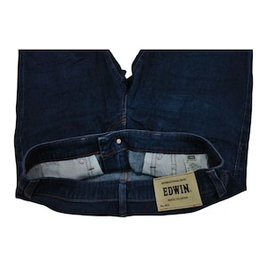 May include: Dark blue denim jeans with a button closure and a visible label. The label reads "EDWIN" and "MADE IN JAPAN". The jeans are folded, showcasing the interior pockets and stitching details. The overall style is classic and casual.