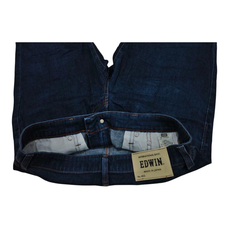 May include: Dark blue denim jeans with a button closure and a visible label. The label reads "EDWIN" and "MADE IN JAPAN". The jeans are folded, showcasing the interior pockets and stitching details. The overall style is classic and casual.