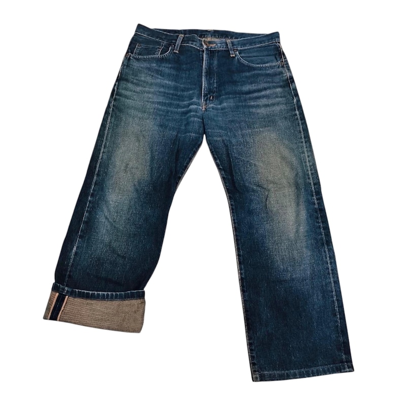 May include: Blue denim jeans with a faded wash. The jeans have a classic five-pocket design, a button fly, and a relaxed fit. The cuffs are rolled up, revealing a contrasting fabric. The jeans are displayed on a white background.