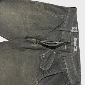 K&ouml;nnte beinhalten: Nahaufnahme einer olivgr&uuml;nen Jeans aus Denim mit wei&szlig;en N&auml;hten. Die Jeans hat einen Rei&szlig;verschluss, mehrere Taschen und einen Knopfverschluss. Die Jeans hat einen Used-Look und ein kleines Etikett mit den Buchstaben "O.U."