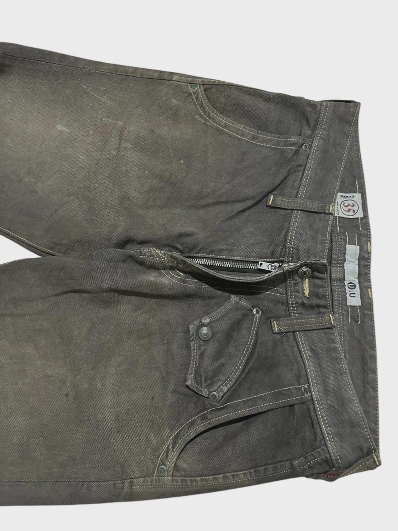 K&ouml;nnte beinhalten: Nahaufnahme einer olivgr&uuml;nen Jeans aus Denim mit wei&szlig;en N&auml;hten. Die Jeans hat einen Rei&szlig;verschluss, mehrere Taschen und einen Knopfverschluss. Die Jeans hat einen Used-Look und ein kleines Etikett mit den Buchstaben "O.U."