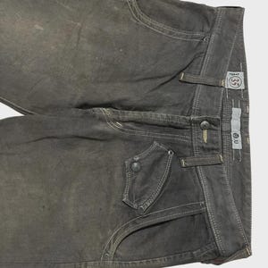 K&ouml;nnte beinhalten: Dunkelgraue Jeans mit wei&szlig;en N&auml;hten. Merkmale sind ein einzigartiges Taschen-Design mit Kn&ouml;pfen, G&uuml;rtelschlaufen sowie Knopf- und Rei&szlig;verschluss. Die Jeans haben ein verwaschenes, abgenutztes Aussehen.