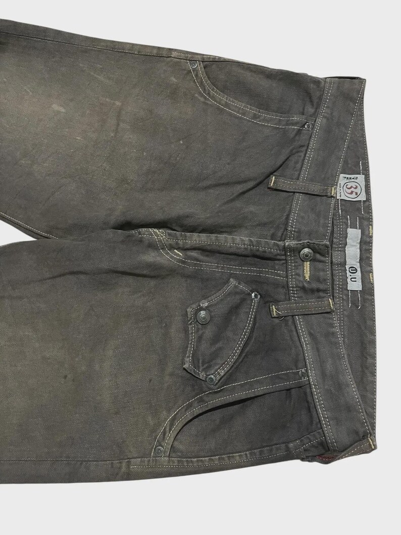 K&ouml;nnte beinhalten: Dunkelgraue Jeans mit wei&szlig;en N&auml;hten. Merkmale sind ein einzigartiges Taschen-Design mit Kn&ouml;pfen, G&uuml;rtelschlaufen sowie Knopf- und Rei&szlig;verschluss. Die Jeans haben ein verwaschenes, abgenutztes Aussehen.