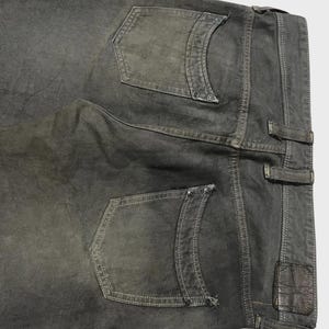 K&ouml;nnte beinhalten: Nahaufnahme der R&uuml;ckseite einer dunkelgrauen Jeans. Die Jeans hat zwei Ges&auml;&szlig;taschen mit dekorativen N&auml;hten und Nieten. Ein kleines Lederpatch ist in der N&auml;he des Bunds sichtbar. Die Jeans ist leicht verblichen, was ihr ein abgenutztes Aussehen verleiht.